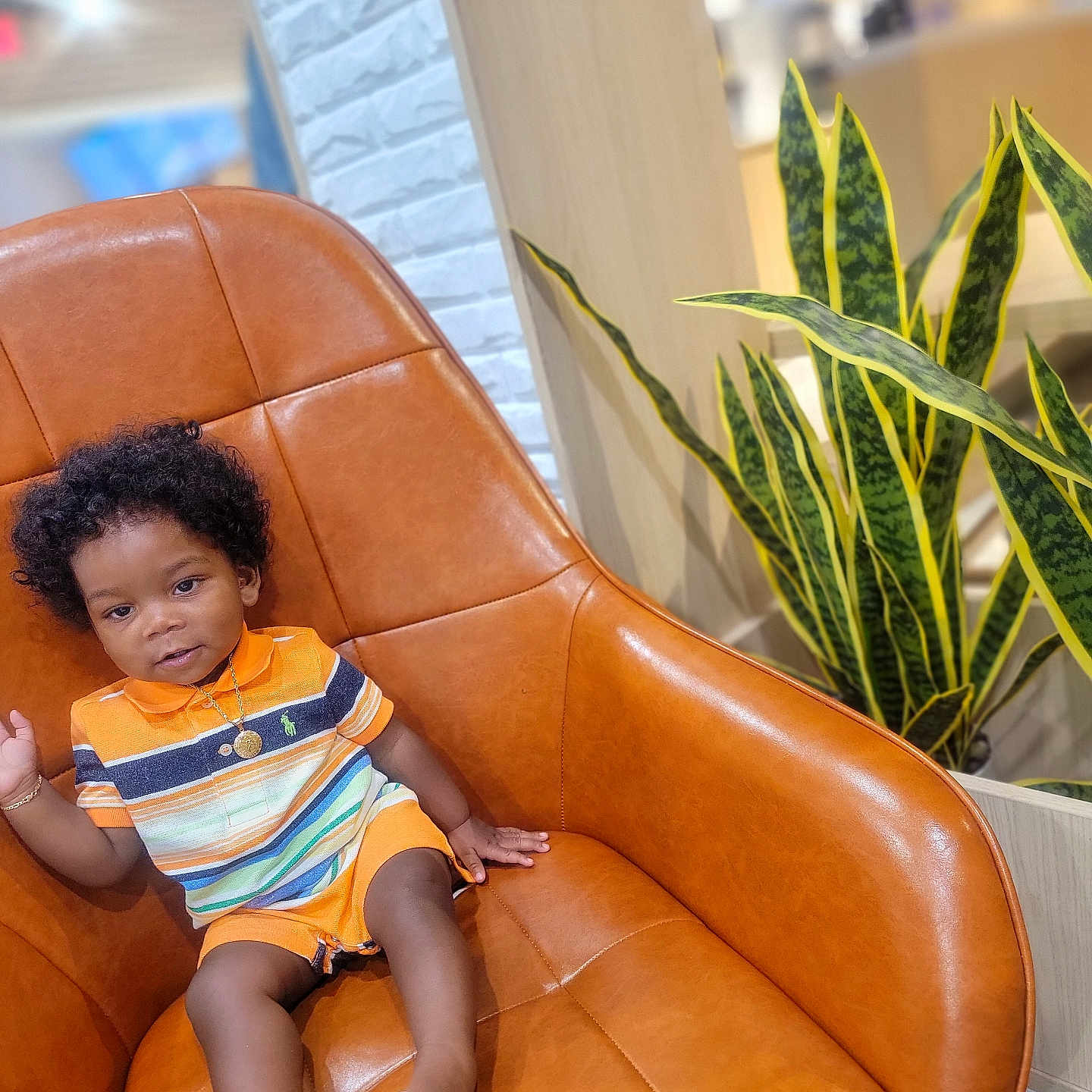 Leoric is registered to the contest to win money with this photo: barefoot, child, curly_hair, cute, gold_bracelet, gold_necklace, green_plant, indoor, leather_chair, modern_interior, orange_outfit, plant, portrait, relaxed, sitting, smiling, striped_clothing, toddler, white_wall, wood_panel