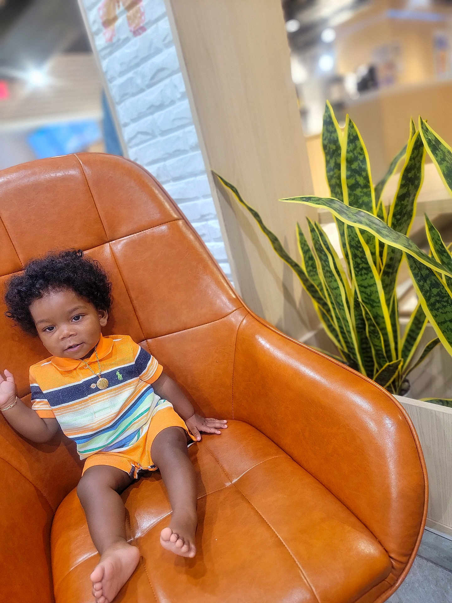 Leoric is registered to the contest to win money with this photo: toddler, child, curly_hair, striped_clothing, orange_outfit, gold_necklace, gold_bracelet, leather_chair, indoor, plant, green_plant, modern_interior, white_wall, wood_panel, barefoot, smiling, sitting, cute, portrait, relaxed