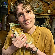 Joey is registered to the contest to win money with this photo: person, kitten, cat, orange_cat, indoor, portrait, smile, holding, wood_paneling, vintage_decor, clock, rifle, cross, couch, pillow, watch, casual_clothing, young_adult, facial_expression, cozy
