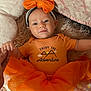 baby, infant, orange_clothing, headband, bow, holding_hand, lying_down, soft_texture, blanket, cute, portrait, child, tutu_skirt, daisy_pattern, smiling, face, person, small_earring, indoor, adorable