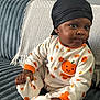 baby, child, onesie, pumpkin, headwrap, black, orange, white, couch, blanket, hand, foot, sitting, indoor, cute, expression, texture, cozy, smile, portrait