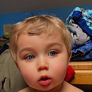 Sebastian is registered to the contest to win money with this photo: toddler, child, face, blond_hair, blue_eyes, indoor, portrait, curious, expression, clothing, pile, furniture, wood, background, skin, person, young_child, closeup, head, hair
