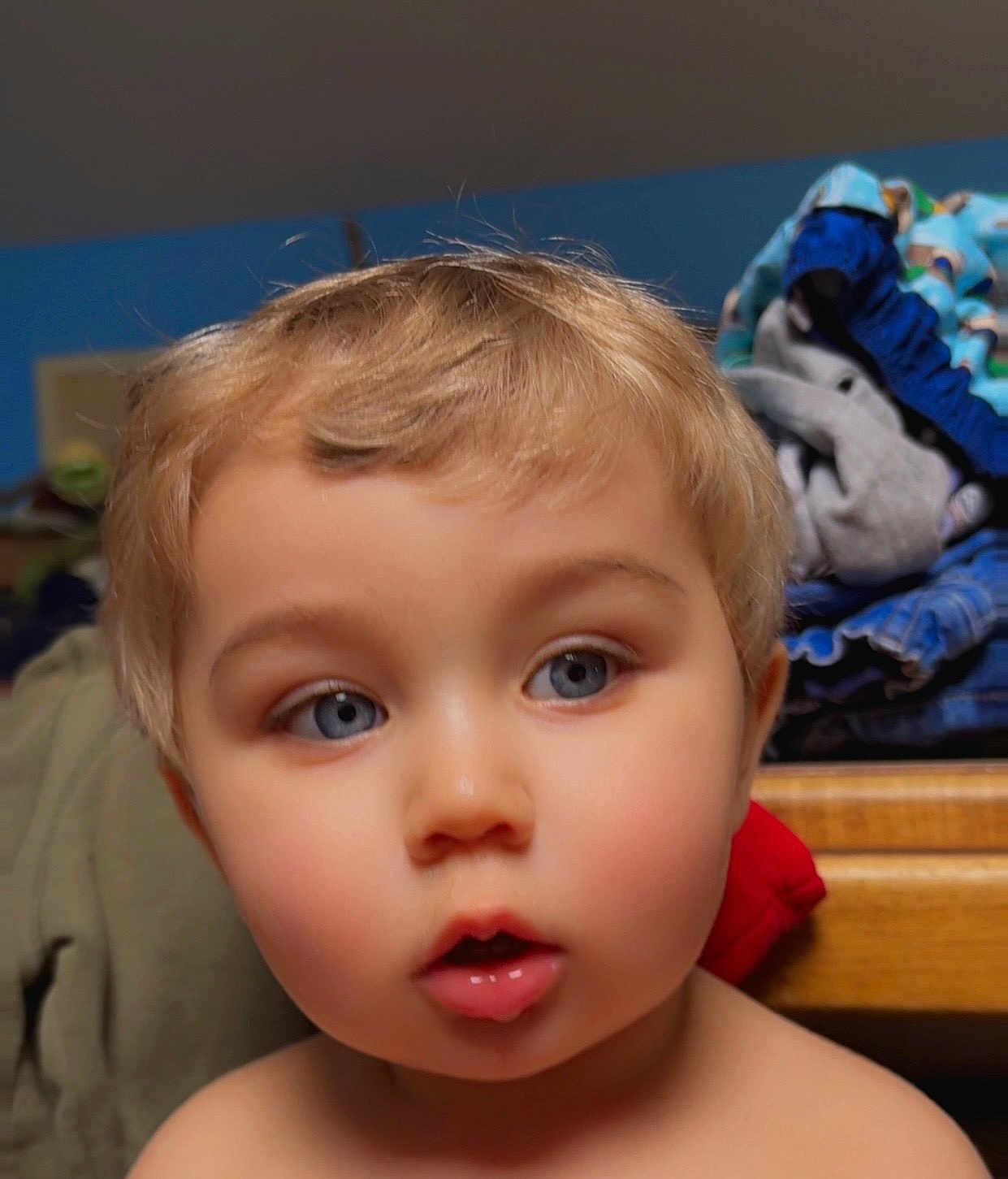 Sebastian is registered to the contest to win money with this photo: toddler, child, face, blond_hair, blue_eyes, indoor, portrait, curious, expression, clothing, pile, furniture, wood, background, skin, person, young_child, closeup, head, hair