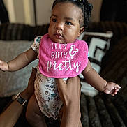 Charm Ross is registered to the contest to win money with this photo: baby, child, infant, person, hands, bib, pink, hair, pigtails, cute, indoor, couch, blurred_background, floral_clothing, skin, face, portrait, holding, expression, love