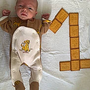 Raphaël a rejoint le concours — aidez-le/la à gagner de superbes lots ! baby, infant, onesie, simba, blanket, biscuit, number_one, food_art, cute, lying_down, celebration, snack, child, portrait, baby_clothing, toddler, light, indoor, soft_texture, playful