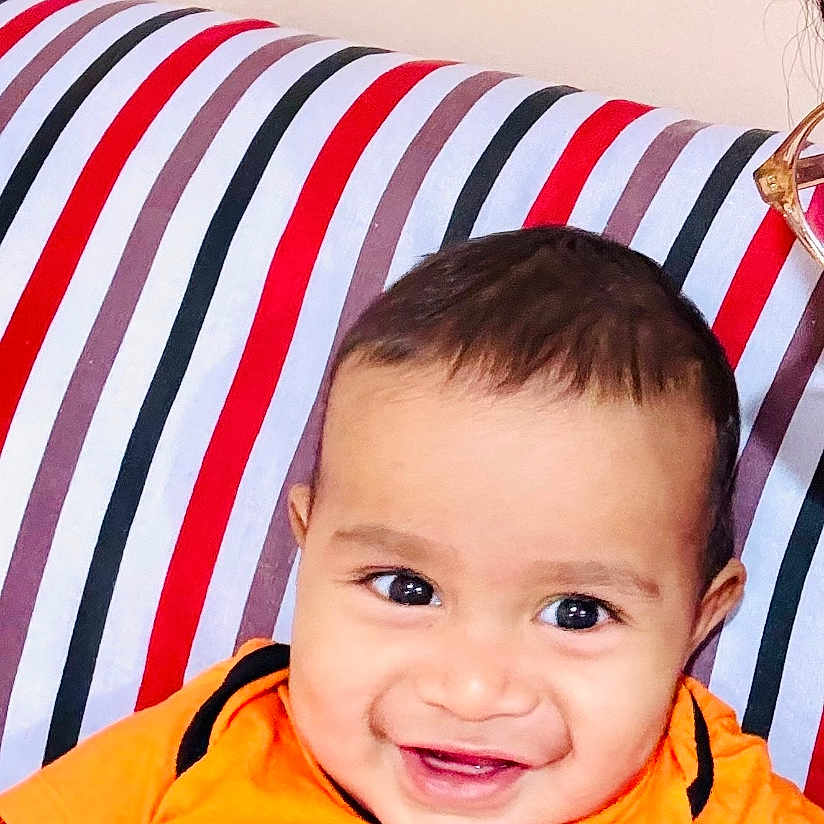 Benaiah is registered to the contest to win money with this photo: baby, bright_colors, cheerful, child, closeup, clothing, cute, face, happy, head, indoor, infant, orange_shirt, person, portrait, seated, skin, smile, striped_couch, young