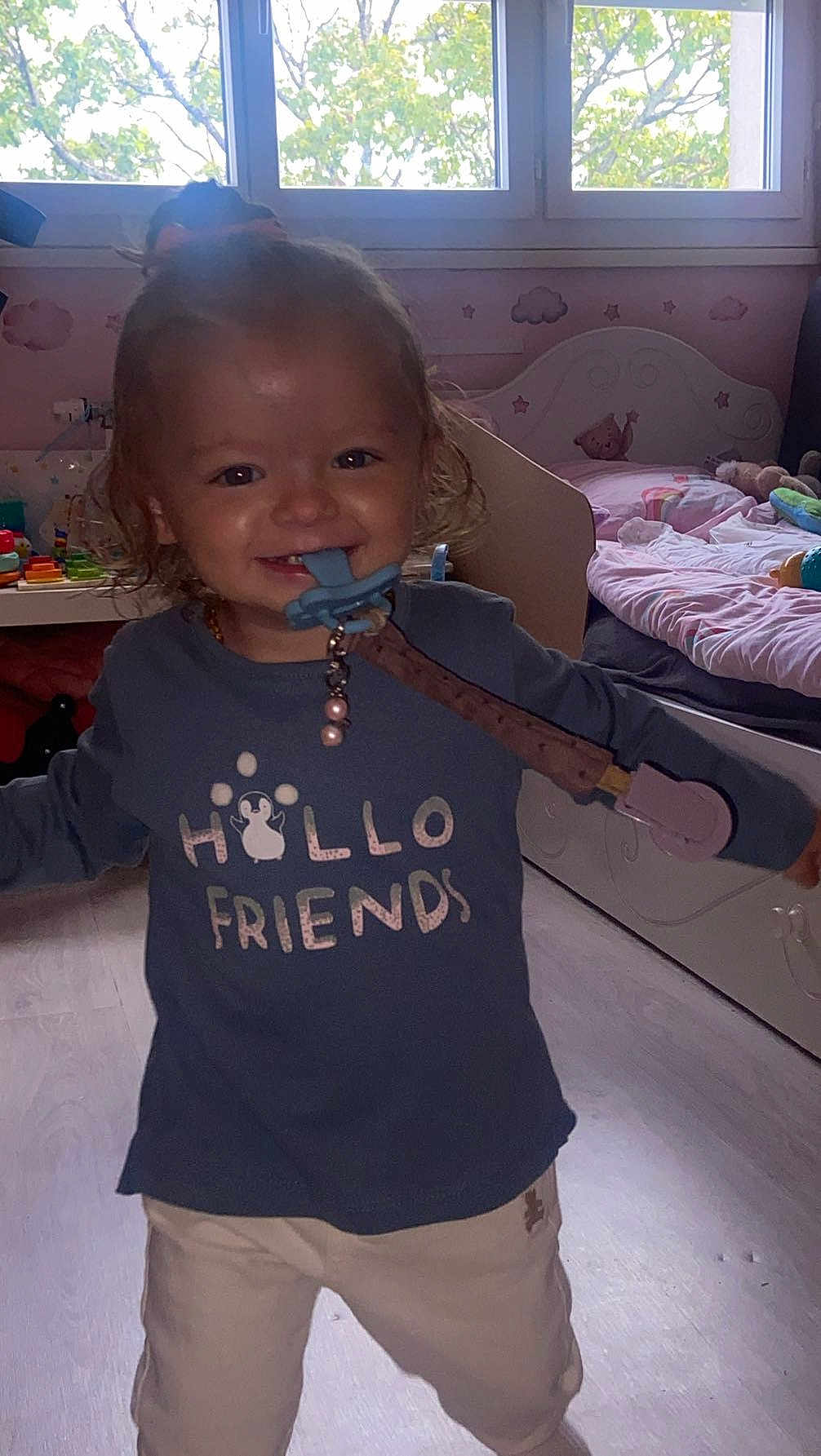 Lyzéa a rejoint le concours — aidez-le/la à gagner de superbes lots ! bed, bedroom, blue_shirt, child, clothing, curly_hair, daylight, floor, happy, hello_friends_text, indoors, pacifier, pants, person, playroom, smiling, toddler, toys, window, young_child