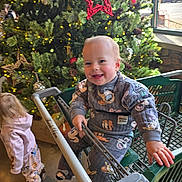 Ellie is registered to the contest to win money with this photo: boots, cart, child, christmas_tree, clothing, festive, floor, holiday_decorations, indoor, lights, pajamas, patterned_pants, person, pine_cones, pink_hoodie, red_star_ornament, shopping_cart, smiling, toddler, window