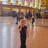 child, indoor, american_flag, people, walking, floor, windows, architecture, public_space, casual_clothing, blonde_hair, black_outfit, shoes, large_hall, light, background, tourists, reflection, crowd, daylight