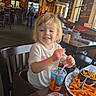 Angel is registered to the contest to win money with this photo: child, toddler, high_chair, restaurant, wood_paneling, drink, straw, sweet_potato_fries, mac_and_cheese, table, chair, person, food, indoor, smiling, blonde_hair, casual_clothing, napkin, fork, dining