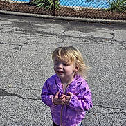 Angel is registered to the contest to win money with this photo: child, blonde_hair, purple_jacket, outdoor, pavement, cracked_road, fence, plants, casual_clothing, young_child, person, standing, daylight, nature, expression, smile, wind, casual_wear, small_child, curly_hair