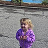 child, blonde_hair, purple_jacket, outdoor, pavement, cracked_road, fence, plants, casual_clothing, young_child, person, standing, daylight, nature, expression, smile, wind, casual_wear, small_child, curly_hair
