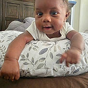 Judah is registered to the contest to win money with this photo: baby, child, cushion, bed, indoor, person, face, arm, hand, expression, curious, pattern, white_clothing, brown_skin, head, lying_down, close_up, room, window, wooden_furniture