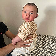 Sacha a rejoint le concours — aidez-le/la à gagner de superbes lots ! baby, child, person, hands, table, pattern, shirt, striped_clothing, pants, indoor, wall, curious, seated, adult_hand, footwear, sitting, expression, skin, head, face