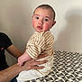 baby, child, person, hands, table, pattern, shirt, striped_clothing, pants, indoor, wall, curious, seated, adult_hand, footwear, sitting, expression, skin, head, face