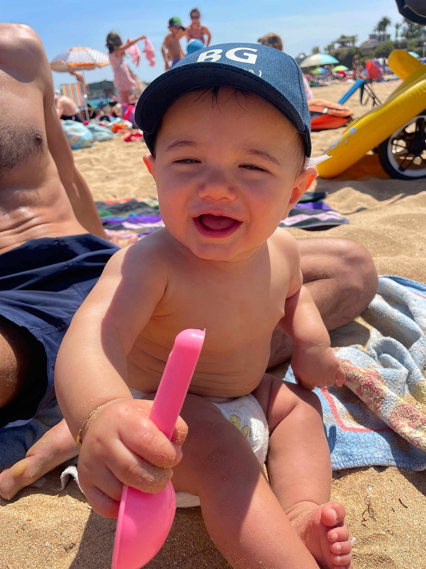 Sacha a rejoint le concours — aidez-le/la à gagner de superbes lots ! baby, child, cap, beach, sand, sunny, smile, toy, shovel, diaper, towel, person, summer, vacation, outdoor, sunlight, happy, play, parent, family