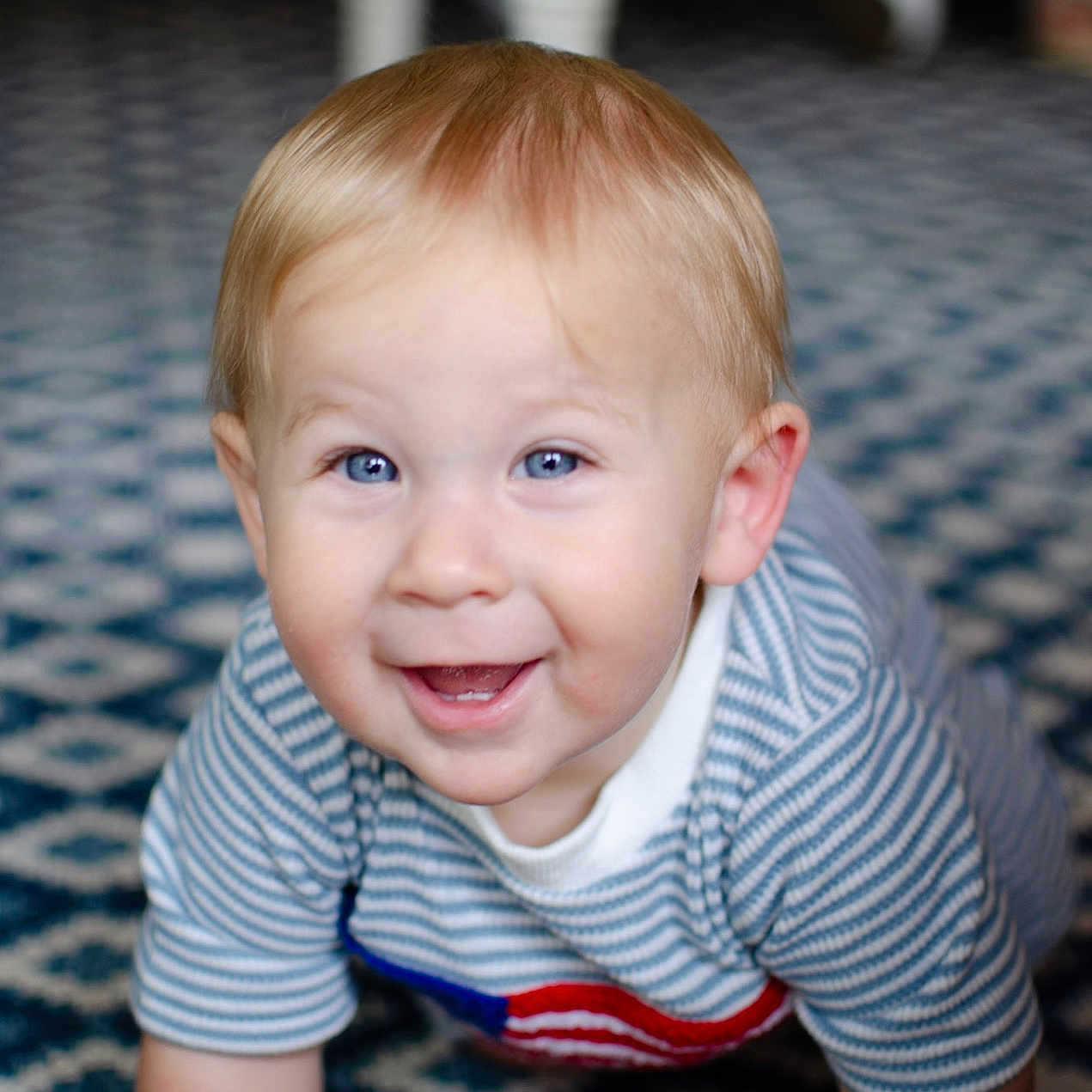 Colson is registered to the contest to win money with this photo: baby, child, crawling, blue_eyes, striped_shirt, carpet, patterned_floor, indoor, smiling, happy, portrait, person, infant, cute, blond_hair, face, young_child, home, floor, toddler