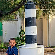 Sohann a rejoint le concours — aidez-le/la à gagner de superbes lots ! black_and_white, blue_clothing, building, child, concrete_base, curious, daylight, garden, lamp_post, lighthouse, nature, outdoor, plants, playful, sandals, short_hair, sitting, striped, toddler, young_child