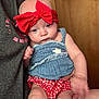 baby, child, red_bow, blue_top, red_checkered_bottoms, indoor, door, person, skin, hand, arm, face, head, clothing, holding, seated, close_up, casual, cute, infant
