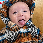 Teddie joined the competition — help win amazing prizes! baby, child, hoodie, fleece, smiling, tongue_out, face, cute, warm_clothing, indoor, table, patterned_clothing, infant, playful, happy, person, portrait, closeup, headwear, cozy