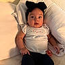 baby, infant, headband, bow, white_top, dark_pants, cushion, pillow, indoors, cute, child, person, portrait, relaxing, looking, wide_eyes, seated, soft_lighting, comfortable, expression