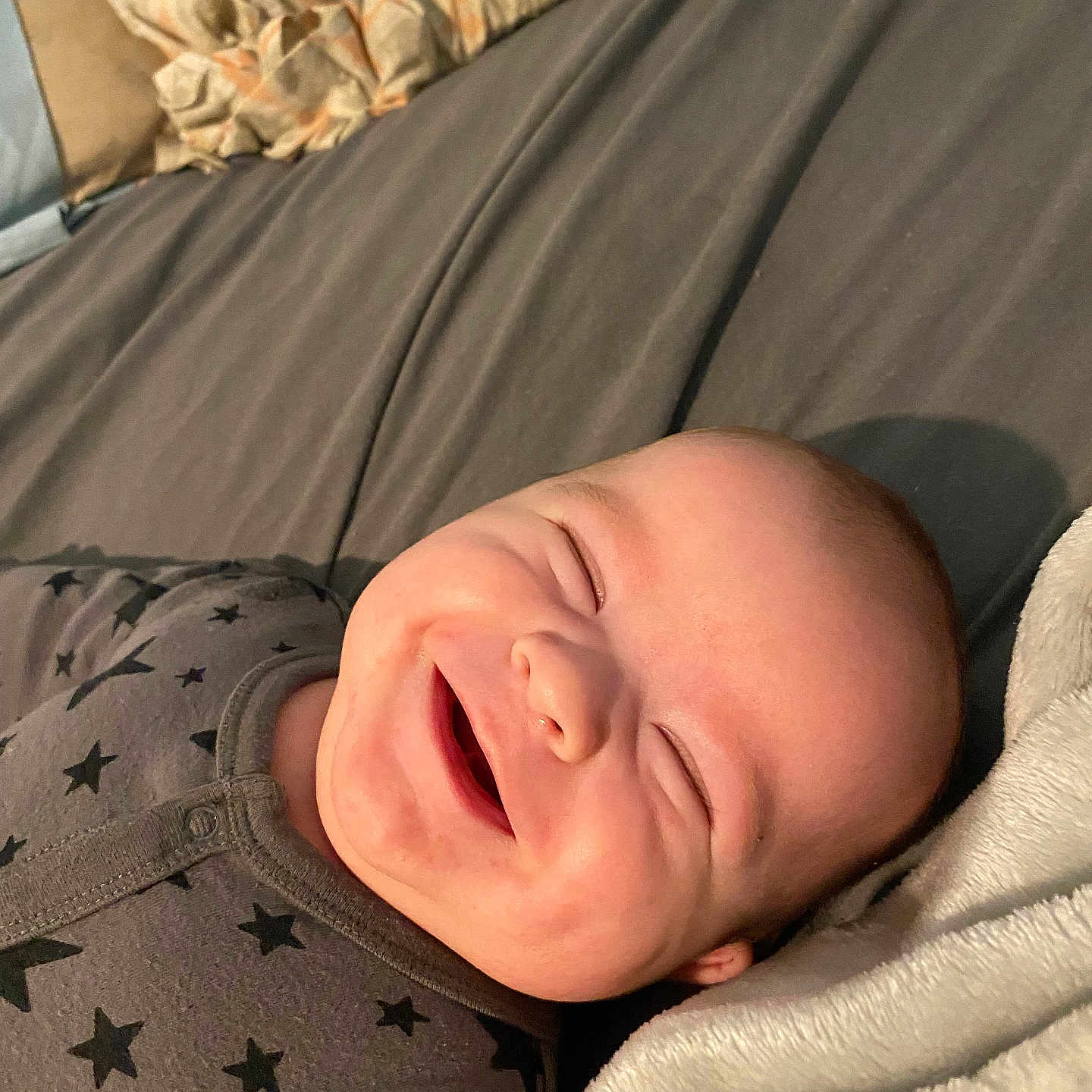 Isaac joined the competition — help win amazing prizes! baby, smiling, onesie, star_pattern, bed, blanket, pillow, face, head, infant, happy, closeup, sleepwear, person, child, cute, cozy, indoor, portrait, resting