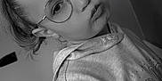 Lou-Ann participe au concours pour gagner de l'argent avec cette photo : child, girl, glasses, pout, hoodie, portrait, black_and_white, indoor, wall, paper_notice, young_person, closeup, hair_tied, eyewear, expression, casual_clothing, face, seated, playful, nostalgic