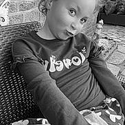 Lou-Ann a rejoint le concours — aidez-le/la à gagner de superbes lots ! child, girl, playful_expression, black_and_white, outdoor, brick_wall, rattan_chair, sweater, patterned_skirt, sitting, portrait, funny_face, garden_gnome, patio, cobblestone, curls, eyes, long_sleeves, casual_clothing, candid