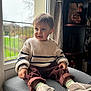 carved_art, casual, child, cozy, curious, cushion, expression, furniture, home, indoor, knitwear, ottoman, pants, portrait, shoes, sitting, soft_lighting, sweater, toddler, window