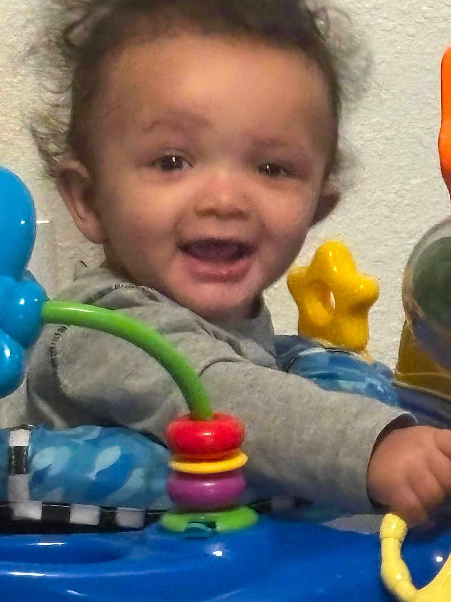 Skyler joined the competition — help win amazing prizes! baby, child, smile, face, toy, colorful_toys, activity_center, indoor, seat, hand, eyes, hair, mouth_open, star_shaped_toy, ring_toy, blue_toy, gray_sweatshirt, portrait, closeup, playtime