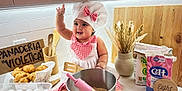 Zoe is registered to the contest to win money with this photo: baby, infant, chef_hat, apron, mixing_bowl, rolling_pin, spatula, eggs, croissant, bread, kitchen, countertop, wooden_spoon, vase, dried_flowers, flour, sugar_box, milestone_sign, smile, playful