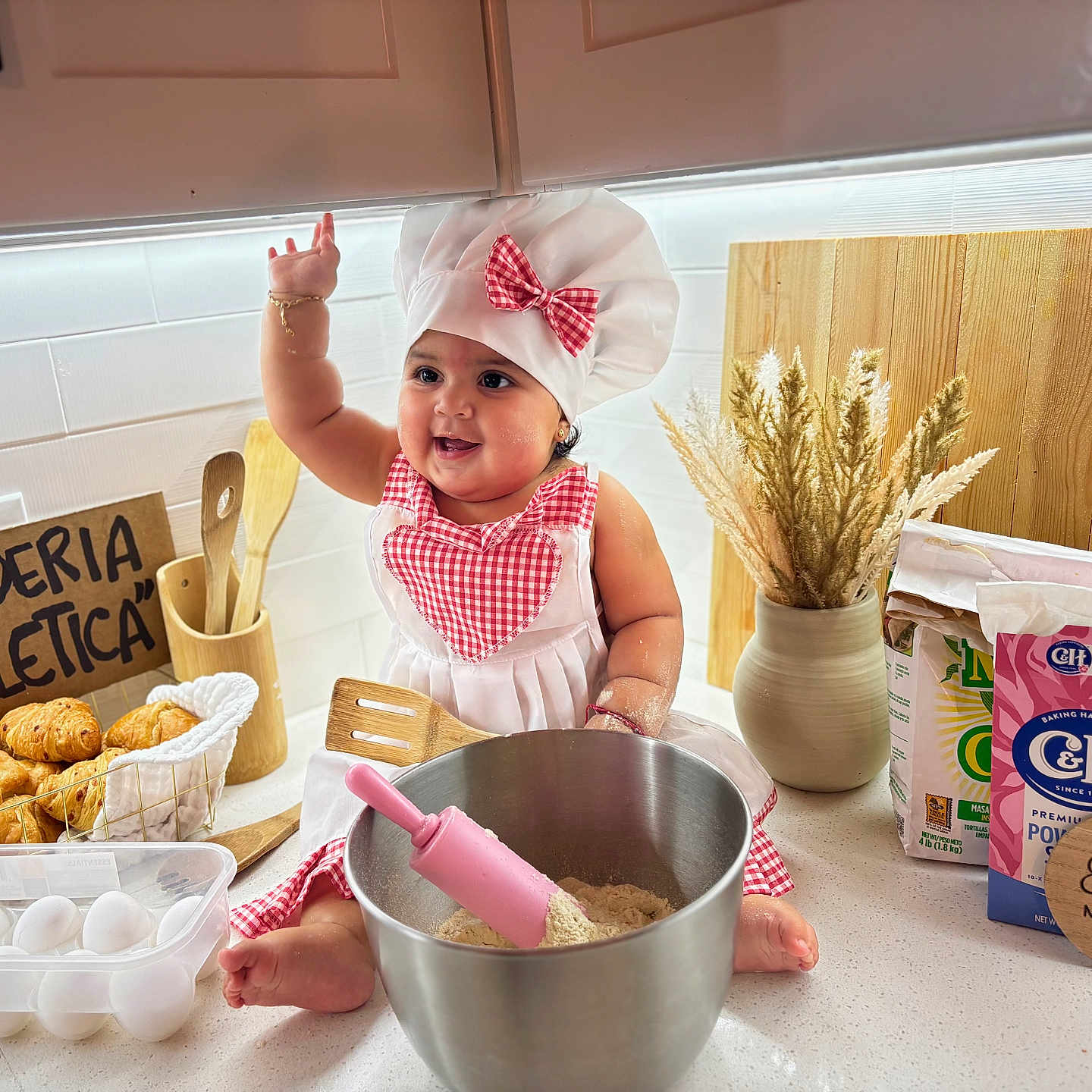 Zoe is registered to the contest to win money with this photo: apron, baby, bread, chef_hat, countertop, croissant, dried_flowers, eggs, flour, infant, kitchen, milestone_sign, mixing_bowl, playful, rolling_pin, smile, spatula, sugar_box, vase, wooden_spoon
