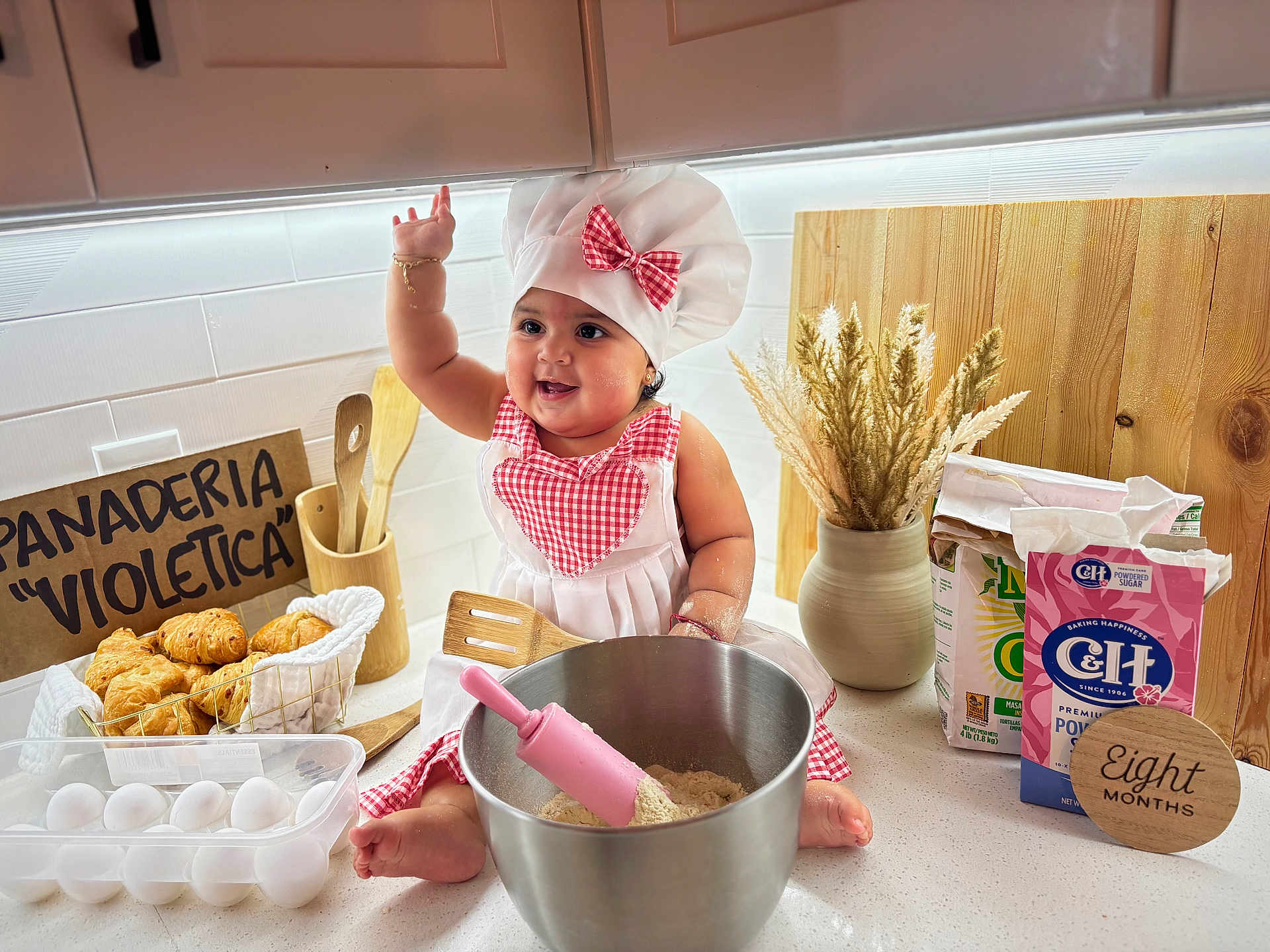 Zoe is registered to the contest to win money with this photo: baby, infant, chef_hat, apron, mixing_bowl, rolling_pin, spatula, eggs, croissant, bread, kitchen, countertop, wooden_spoon, vase, dried_flowers, flour, sugar_box, milestone_sign, smile, playful