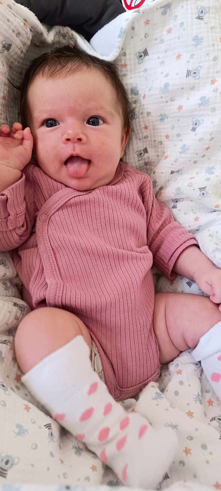 Louna a rejoint le concours — aidez-le/la à gagner de superbes lots ! baby, infant, tongue_out, pink_onesie, white_socks, polka_dots, blanket, koala_pattern, stars, cute, playful, lying_down, soft_texture, child, smiling, cozy, indoor, portrait, skin, hand