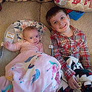 Willie is registered to the contest to win money with this photo: baby, child, siblings, blanket, unicorn, plaid_shirt, couch, pillow, smiling, happy, indoors, face, person, clothing, infant, toddler, young_child, home, blanket_pattern, relaxing