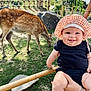 baby, child, smiling, outdoor, deer, grass, rocks, bamboo_fence, hat, bonnet, person, nature, animal, happy, greenery, sunlight, portrait, holding, cute, tongue_out