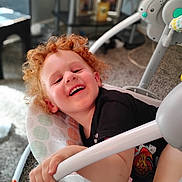 Kyrie is registered to the contest to win money with this photo: child, toddler, curly_hair, smiling, laughing, baby_swing, indoor, carpet, black_shirt, hand, toy, furniture, home, playful, happy, person, seat, grip, closeup, young