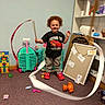 toddler, child, curly_hair, smiling, ribbon, toy, carpet, indoor, playroom, project_board, shelf, plastic_toy, red_shoes, black_tshirt, grey_pants, happy, young_child, playing, motion_blur, colorful