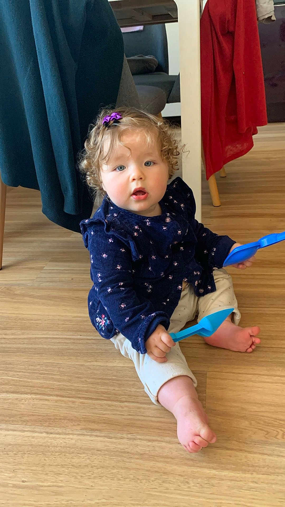 Louise a rejoint le concours — aidez-le/la à gagner de superbes lots ! toddler, child, curly_hair, hair_clip, blue_shovel, plastic_toy, sitting, wooden_floor, indoor, clothing, pants, long_sleeve, purple, red_cloth, chair, foot, hand, face, expression, home