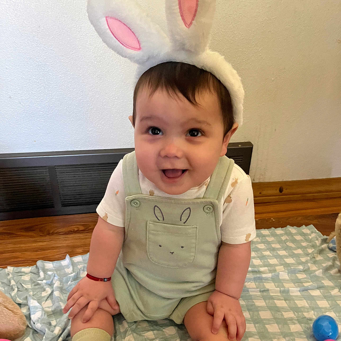 Lorenzo is registered to the contest to win money with this photo: baby, bunny_ears, smiling, child, indoor, floor, blanket, plastic_eggs, green_overall, cute, happy, playtime, toy, sock, wooden_floor, face, person, easter, holiday, candid