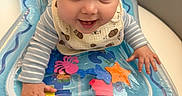 Lorenzo is registered to the contest to win money with this photo: baby, infant, smiling, face, hands, inflatable_toy, water_toy, starfish, crab, blue, striped_clothing, bib, cute, indoor, playtime, child, person, happy, closeup, toddler