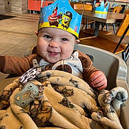 Lorenzo is registered to the contest to win money with this photo: baby, high_chair, blanket, pacifier, crown, smile, face, child, indoor, restaurant, table, chair, wood, toy, clothing, happy, cute, infant, portrait, playful
