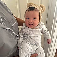 William is registered to the contest to win money with this photo: baby, child, infant, person, hat, knit_hat, bear_ears, smile, happy, holding, adult, clothing, onesie, indoor, door, wood_floor, face, hand, cute, portrait