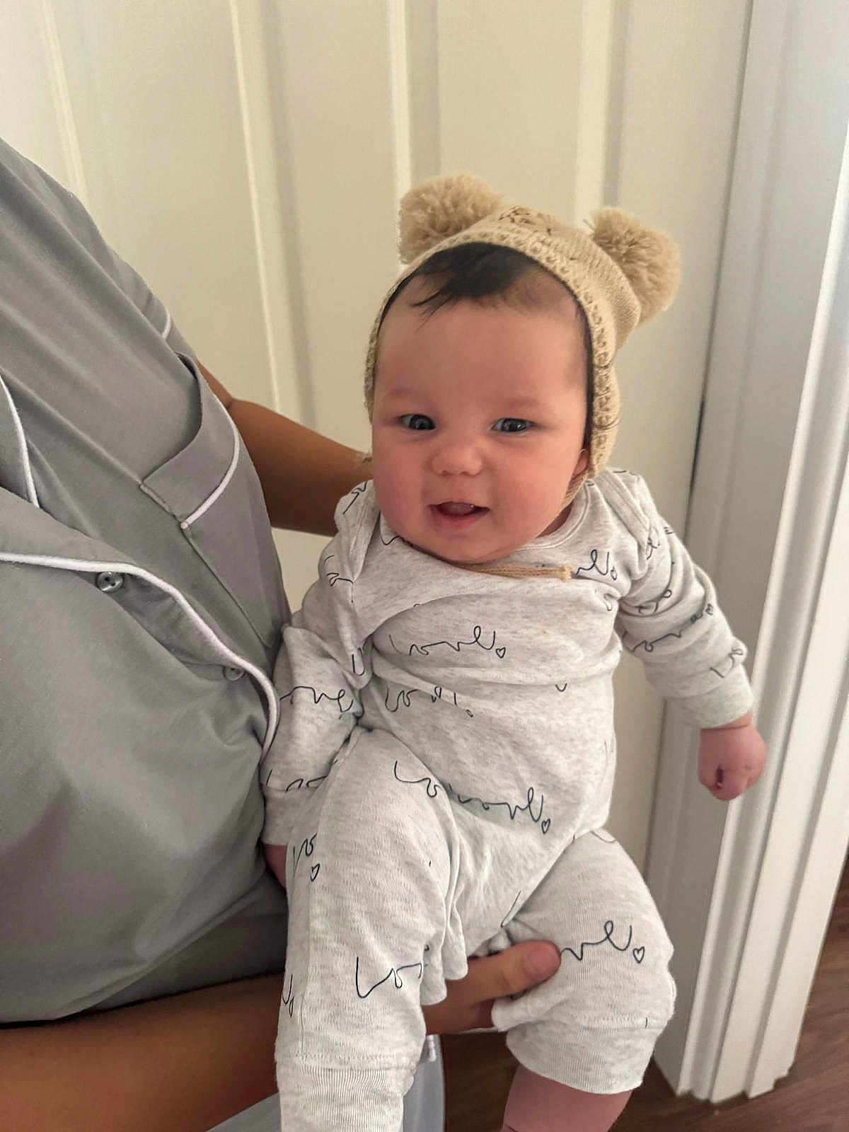William is registered to the contest to win money with this photo: baby, child, infant, person, hat, knit_hat, bear_ears, smile, happy, holding, adult, clothing, onesie, indoor, door, wood_floor, face, hand, cute, portrait