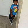 Precise is registered to the contest to win money with this photo: black_pants, blue_shirt, cap, cartoon_shirt, child, earrings, fashion, floor, full_body, hallway, home, indoors, necklace, portrait, serious_expression, sneakers, standing, tile_floor, toddler, wall
