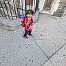 backpack, building, casual_clothing, child, daylight, iron_railings, knit_cap, outdoor, pavement, plaid_shirt, portrait, red_shoes, sidewalk, snack, standing, street, toddler, urban, walking, wide_angle