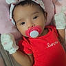 baby, baby_clothing, bed, blanket, bow, cartoon, child, close_up, cute, face, headband, indoors, infant, lying_down, mittens, pacifier, pink_bedding, portrait, red_outfit, smiling