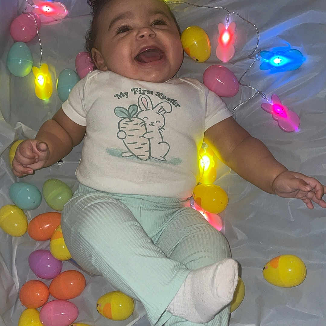 Normani joined the competition — help win amazing prizes! baby, child, easter_eggs, string_lights, bunny_lights, smiling, happy, infant, white_background, colorful, festive, holiday, celebration, clothing, pants, socks, lying_down, cute, indoors, joyful