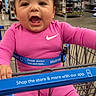 baby, child, shopping_cart, pink_clothing, smiling, happy, indoor, store, retail, aisle, walmart, seat_belt, app_promotion, curly_hair, person, retail_environment, shopping, infant, clothing, safety