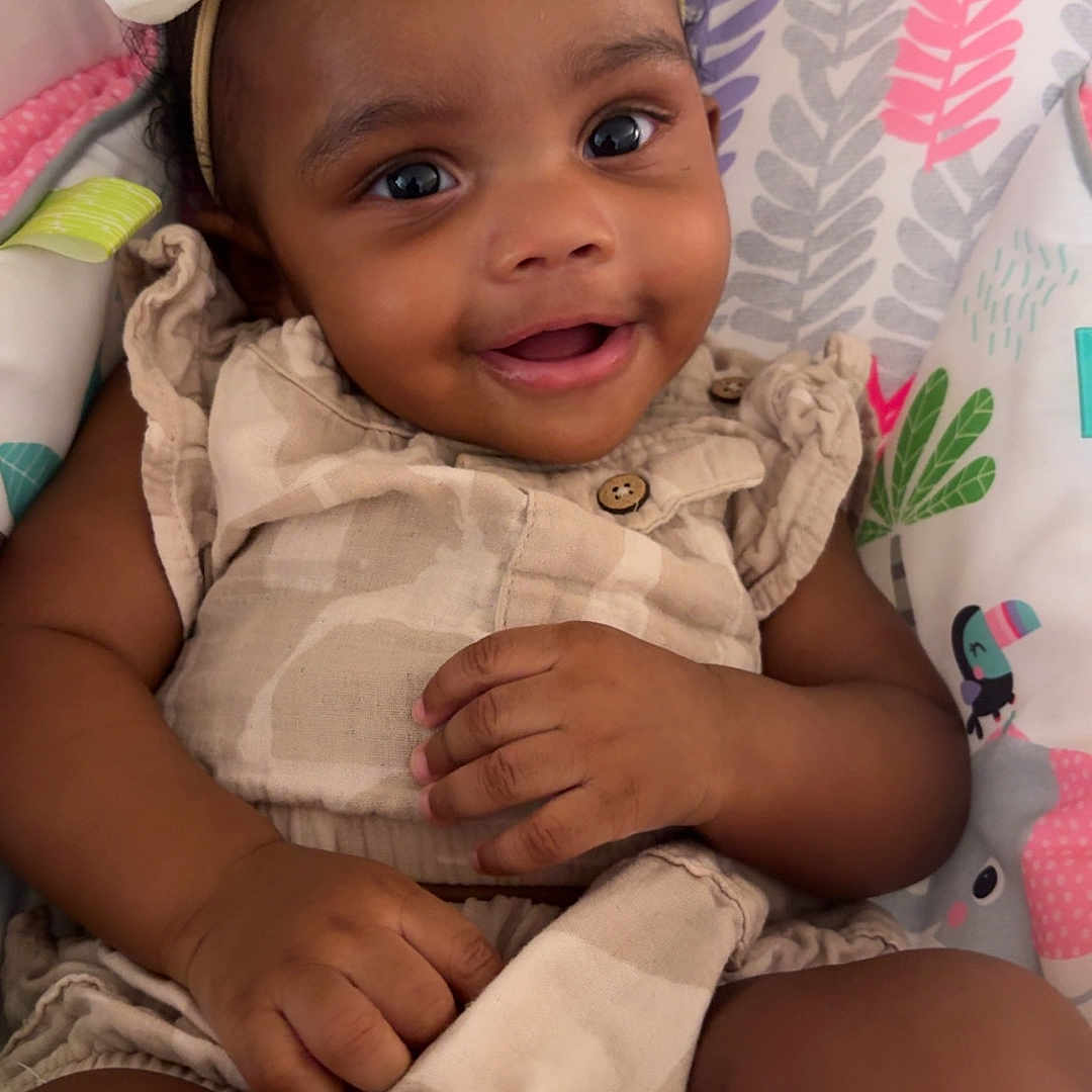 Saniyah is registered to the contest to win money with this photo: baby, child, infant, smiling, portrait, bow, headband, dress, sitting, lying_down, blanket, playmat, patterned_background, indoor, face, hands, legs, happy, cute, photography